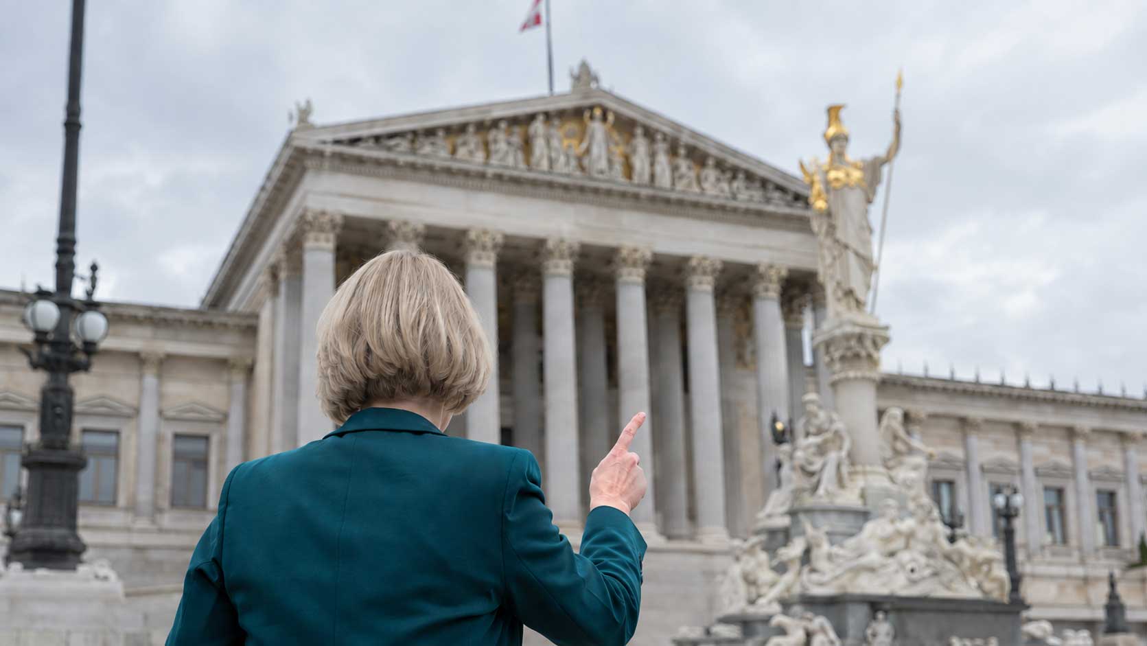 mikl-leitern-lohnnebenkosten Frau von hinten mit blondem Bob und dunkelgrünem Blazer, erhobener Zeigefinger, steht vor dem österreichischen Parlament in Wien mit der goldenen Pallas-Athene-Statue